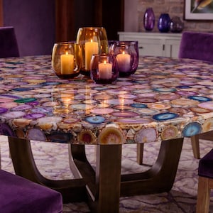 May include: A round dining table with a colorful agate stone top. The table features a dark wooden base and is surrounded by purple velvet chairs. Glass candle holders with lit candles are on the table.