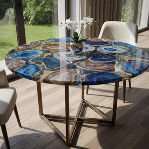 Puede incluir: Mesa de comedor redonda con una tapa de piedra de ágata pulida con patrones azules, marrones y blancos. La mesa tiene una base de metal color bronce y está rodeada de sillas de tonos neutros. Un jarrón con flores blancas está sobre la mesa.