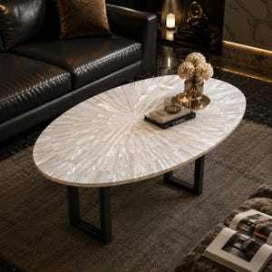 May include: An oval coffee table featuring a tabletop inlaid with iridescent white mother-of-pearl tiles arranged in a sunburst pattern. The table is supported by two black metal U-shaped legs. A book with the word CHANEL is placed on the surface.