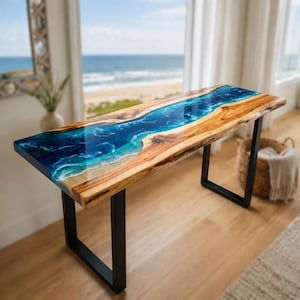Op de afbeelding: Een rechthoekige houten tafel met een blauw epoxyhars rivierontwerp. De tafel heeft een natuurlijke houtnerf en zwarte metalen poten. De hars simuleert oceaangolven en een zandstrand. De tafel staat voor een raam met uitzicht op zee.