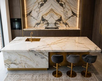 May include: A contemporary kitchen with a marble island and matching splashback. The island has a gold tap and three grey velvet bar stools. Dark wood cabinets and a patterned rug complete the luxurious design.