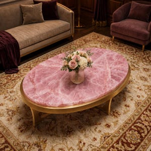 May include: Oval coffee table with a pink, marbled surface and a gold-colored metal frame. A vase of flowers sits on the table, which is placed on a patterned rug. The table is a focal point in a living room setting.
