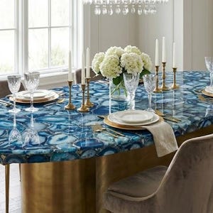 Puede incluir: Una mesa de comedor ovalada con una tapa de piedra de ágata azul, base de latón y patas doradas. La mesa está puesta con platos blancos, cubiertos dorados y copas de vino. Flores blancas y velas adornan la mesa.