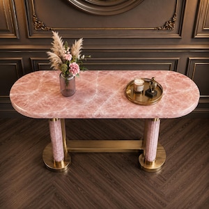 May include: A pink marble-topped console table with a gold-coloured base and legs. The table features a vase of flowers and a decorative tray with a candle and figurine. The table is 152 cm long.