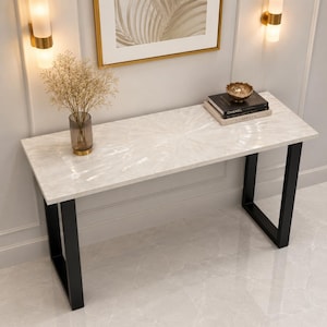 Può includere: Un tavolo console rettangolare con un piano bianco a motivo radiale e due gambe in metallo nero a forma di U. La superficie del tavolo ospita un vaso in vetro trasparente con fiori secchi, due libri neri impilati e una piccola ciotola dorata.