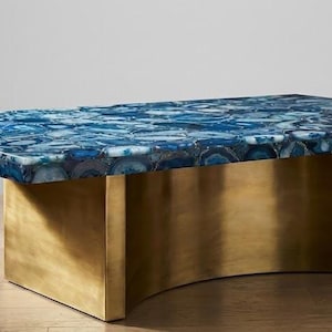 Puede incluir: Una mesa de centro rectangular con una tapa de piedra de ágata azul y una base de metal dorado. La parte superior de la mesa es un mosaico de patrones azules y blancos. La base tiene un diseño curvo.