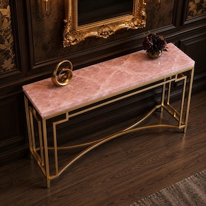 May include: A rectangular console table with a pink marble top and a gold-colored metal frame. The frame features geometric designs on the sides and a curved support beam. A gold sculpture and a vase of flowers sit on the table.