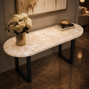 May include: An oval console table with a translucent, cream-coloured stone top and black metal U-shaped legs. A gold-toned vase holds a bouquet of white roses, and two stacked books titled CHANEL and KINFOLK sit nearby with a gold bowl.