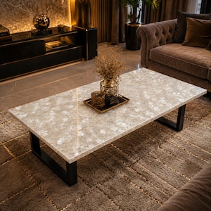 May include: A rectangular coffee table featuring a polished light beige stone top with a mottled white pattern and a smooth finish. The table is supported by two sturdy black metal U-shaped legs, providing a stable base for the heavy stone surface.