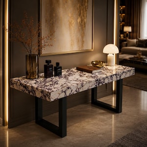 Può includere: Un tavolo console rettangolare con piano in marmo bianco e viola scuro, sostenuto da due gambe in metallo nero a forma di U. La superficie ospita un alto vaso in vetro ambrato con rami, due flaconi scuri, un taccuino marrone e una ciotola dorata.