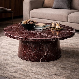 Puede incluir: Una mesa de centro redonda con una tapa de mármol burdeos y una base cilíndrica. La mesa tiene un patrón veteado rojo oscuro y blanco. Un cuenco negro, libros y velas están sobre la mesa. La mesa está sobre una alfombra gris.