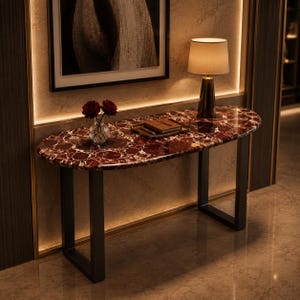 May include: An oval console table featuring a deep red marble top with white veining and two black metal U-shaped legs. On the surface sits a clear glass vase with two dark red roses, a rectangular tray, and a small lamp with a beige shade.
