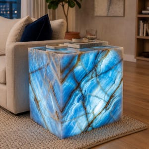 May include: A cube-shaped side table with a striking blue and brown veined pattern. The table is illuminated from within, creating a glowing effect. The top surface holds a book, a small dish, and a glass. The text "STONE & LIGHT" is visible on the book.