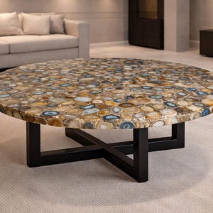 May include: A circular coffee table featuring a mosaic surface of polished stones in brown, beige, and blue hues. It stands on a black metal base with an X-shaped design. The table is a statement piece in a contemporary living space.