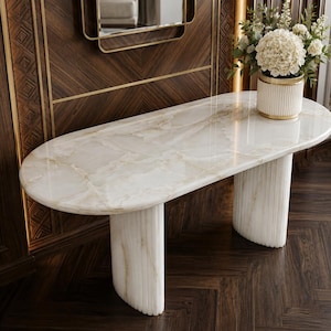 May include: A marble console table with an oval top and fluted legs. The table has a cream and beige colour scheme, with a polished surface. A vase of white flowers sits on the table. The table is set against a dark wood panel wall.