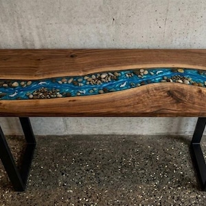 Op de afbeelding: Een houten riviertafel met een donkere walnoot afwerking. Het middenstuk heeft een blauwe epoxyhars rivier met ingebedde stenen en witte golfdetails. De tafel heeft zwarte metalen poten.
