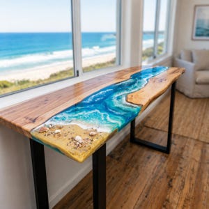 May include: A wooden console table with a resin river design. The resin is blue and white, mimicking ocean waves, with a sandy beach effect at the edge. The table has black metal legs and is near a window with an ocean view.
