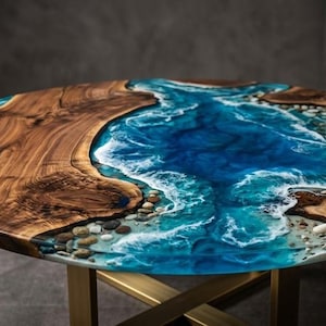 May include: A round coffee table with a unique design. The tabletop features a combination of natural wood and a blue epoxy resin that mimics the ocean. The table is supported by a gold-colored metal base.