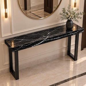 May include: A long rectangular console table featuring a polished black marble top with white veining. The table is supported by two sturdy black metal U-shaped legs. A dark ribbed vase containing green foliage sits on the right side of the surface.
