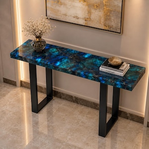 Può includere: Un tavolo console rettangolare con un piano in pietra lucida blu e nera dal vivace motivo marmorizzato. Il tavolo è sostenuto da due gambe in metallo nero a forma di U. Un vaso di vetro con fiori bianchi e dei libri poggiano sulla superficie.