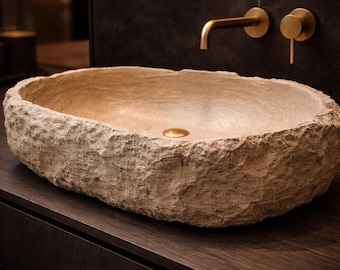 May include: A natural stone vessel sink with a rough, textured exterior and a smooth interior. The sink is a light beige color and has a gold-colored drain. Gold-colored faucet fixtures are mounted on the dark wall behind the sink.