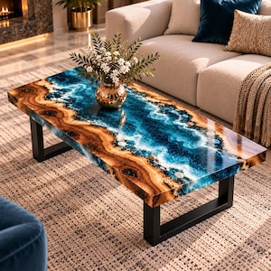 Peut inclure: Une table basse rectangulaire avec un bord en bois naturel et un design de rivière central en résine bleue incrustée de pierres blanches. La table repose sur deux pieds en métal noir en forme de U et porte un vase doré avec des fleurs blanches.