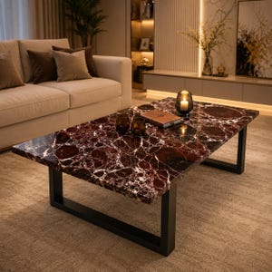May include: A rectangular coffee table featuring a polished dark red marble top with prominent white veining. The table is supported by a sturdy black metal frame with two U-shaped legs. A small candle holder and a closed book sit on the surface.