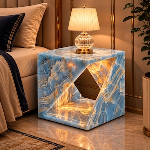 Puede incluir: Una mesa auxiliar con vetas de mármol azul y dorado con un diseño geométrico recortado. Una lámpara con una pantalla plisada y una base de cristal se encuentra encima. La mesa está iluminada desde dentro, creando un brillo cálido. La mesa está al lado de una cama.