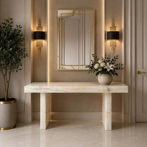 May include: An elegant hallway scene with a marble console table, a gold-framed mirror, and wall lights with black shades. A vase of white flowers is on the table. A potted plant is on the left, and a door is on the right.
