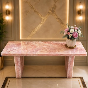 May include: A rectangular pink onyx table with a matching base. The table is topped with a bouquet of pink and white flowers in a striped vase. The table is in a well-lit room with a decorative wall.
