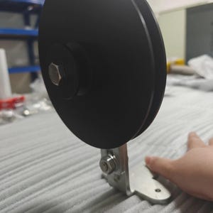 Könnte beinhalten: Eine schwarze, kreisförmige Riemenscheibe, die auf einem silbernen, L-förmigen Metallhalter montiert ist. Die Riemenscheibe verfügt über eine zentrale Sechskantschraube und einen gerillten Außenrand. Der Halter hat eine flache Basis mit vorgebohrten Löchern.