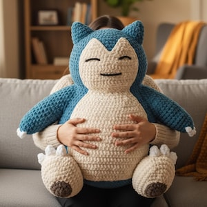 Puede incluir: Un gran peluche de Snorlax hecho a mano con ganchillo. El juguete tiene una cabeza y brazos azules, un cuerpo color crema y detalles blancos y marrones en los pies. El Snorlax está siendo sostenido por una persona.