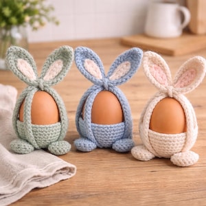 Op de afbeelding: Drie handgemaakte gehaakte eierhouders in pastelkleuren: groen, blauw en crème. Elke houder heeft de vorm van een konijn, met lange oren en een zakje om een ei in te plaatsen. De eieren zijn bruin en contrasteren met de zachte kleuren.