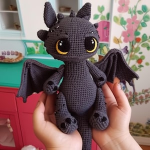 May include: A handmade, dark gray crochet dragon with large, expressive eyes featuring yellow irises. The dragon has wings, small horns, and paw-like feet. The plush toy is held in a person's hands.