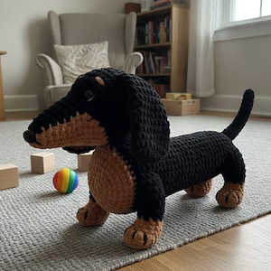 Puede incluir: Un animal de peluche de perro salchicha tejido a ganchillo con cuerpo, cabeza y orejas largas de color negro, con detalles en color canela en el hocico, el pecho y las patas. El juguete tiene una nariz negra pequeña y ojos oscuros.