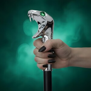 May include: A walking stick featuring a polished silver-tone handle shaped like a cobra head with its mouth open, sharp fangs, and a single vibrant green gemstone eye. The handle has a textured, studded surface and connects to a smooth black shaft.