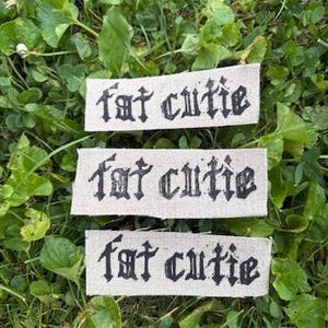 May include: Three rectangular fabric patches made of light beige linen material. Each patch features the text "fat cutie" printed in a bold, black, gothic-style font. The patches are arranged horizontally, one above the other, in a stacked formation.