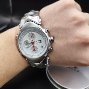 Puede incluir: Un reloj de pulsera Oakley plateado con esfera blanca y detalles rojos. El reloj presenta m&uacute;ltiples esferas, una pantalla de fecha y una correa plateada. El reloj se muestra en una mu&ntilde;eca.