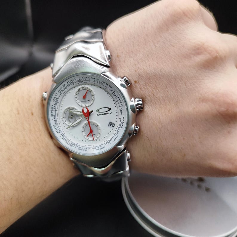 Puede incluir: Un reloj de pulsera Oakley plateado con esfera blanca y detalles rojos. El reloj presenta m&uacute;ltiples esferas, una pantalla de fecha y una correa plateada. El reloj se muestra en una mu&ntilde;eca.