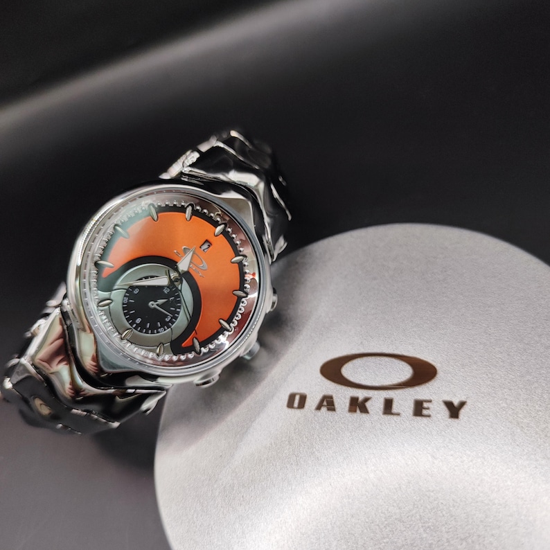 Pu&ograve; includere: Un orologio da polso Oakley argentato con quadrante arancione e nero. L'orologio ha un cinturino unico e strutturato ed &egrave; posto su una superficie grigia accanto a una custodia grigia con il logo Oakley.
