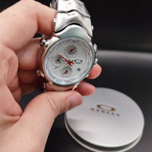 Puede incluir: Un reloj Oakley plateado con esfera blanca y detalles rojos. El reloj presenta m&uacute;ltiples subesferas y una pantalla de fecha. El reloj se sostiene en una mano, con una caja plateada de la marca Oakley en el fondo.