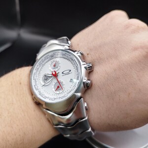 Puede incluir: Un reloj de pulsera Oakley plateado con esfera blanca, m&uacute;ltiples subesferas y agujas rojas. El reloj tiene una pantalla de fecha y una correa texturizada &uacute;nica. El reloj est&aacute; en una mu&ntilde;eca.