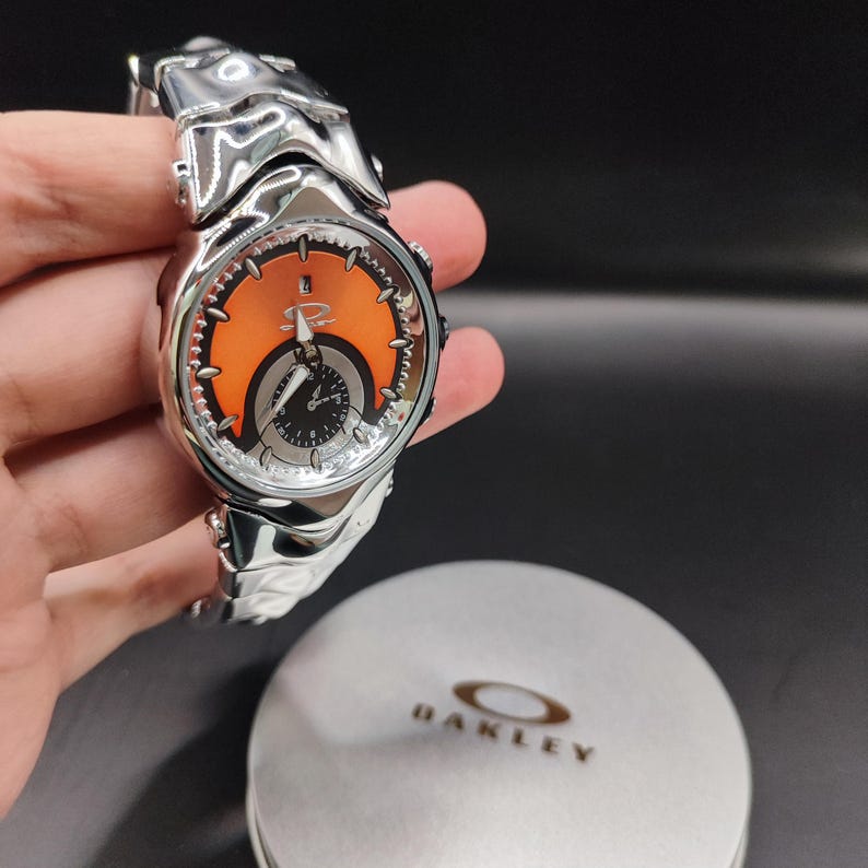 Pu&ograve; includere: Un orologio Oakley color argento con quadrante arancione e nero. L'orologio ha un cinturino scolpito unico ed &egrave; esposto con una custodia Oakley argentata.