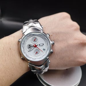 Puede incluir: Un reloj de pulsera Oakley plateado con esfera blanca y detalles rojos. El reloj presenta m&uacute;ltiples esferas y una pantalla de fecha. La correa tambi&eacute;n es plateada y tiene un dise&ntilde;o esculpido &uacute;nico. El reloj se muestra en una mu&ntilde;eca.