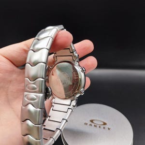 Puede incluir: Un reloj Oakley plateado con una correa de acero inoxidable. La esfera del reloj es visible, junto con la parte trasera del reloj, que tiene texto y un logotipo. El reloj se sostiene en una mano, con la caja Oakley en el fondo.