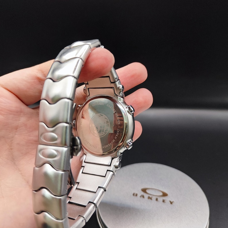 Puede incluir: Un reloj Oakley plateado con una correa de acero inoxidable. La esfera del reloj es visible, junto con la parte trasera del reloj, que tiene texto y un logotipo. El reloj se sostiene en una mano, con la caja Oakley en el fondo.