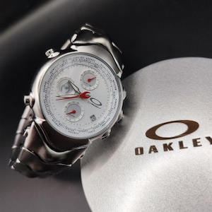 May include: A silver Oakley watch with a white face, red hands, and multiple subdials. The watch features a stainless steel band and is displayed next to a gray Oakley box with the brand's logo.