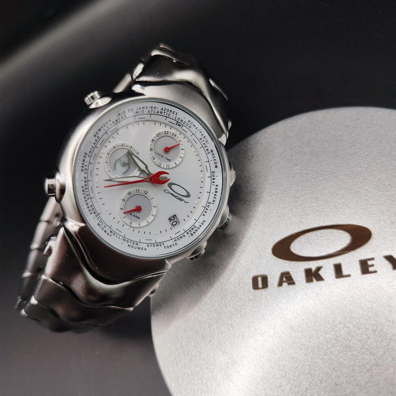 Puede incluir: Un reloj Oakley plateado con esfera blanca, manecillas rojas y m&uacute;ltiples subesferas. El reloj presenta una correa de acero inoxidable y se muestra junto a una caja gris Oakley con el logotipo de la marca.