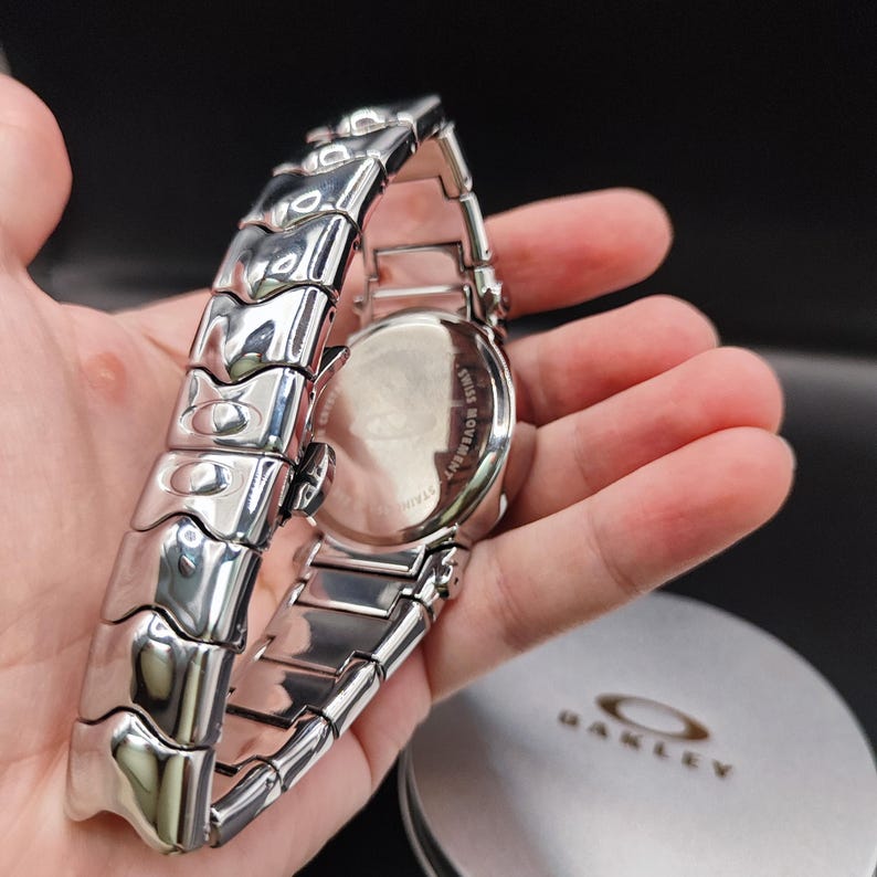Pu&ograve; includere: Un orologio Oakley argentato con retro in acciaio inossidabile e bracciale abbinato. L'orologio ha un quadrante rotondo e una finitura lucida. L'orologio &egrave; mostrato con la sua custodia originale.