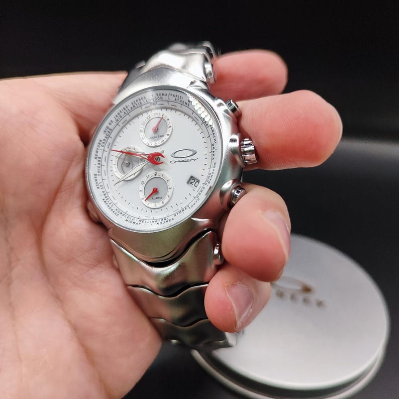 Puede incluir: Reloj de pulsera Oakley plateado con esfera blanca y detalles rojos. El reloj presenta m&uacute;ltiples subesferas, una pantalla de fecha y una correa de metal texturizado. El reloj se sostiene en una mano, mostrando su dise&ntilde;o y detalles.